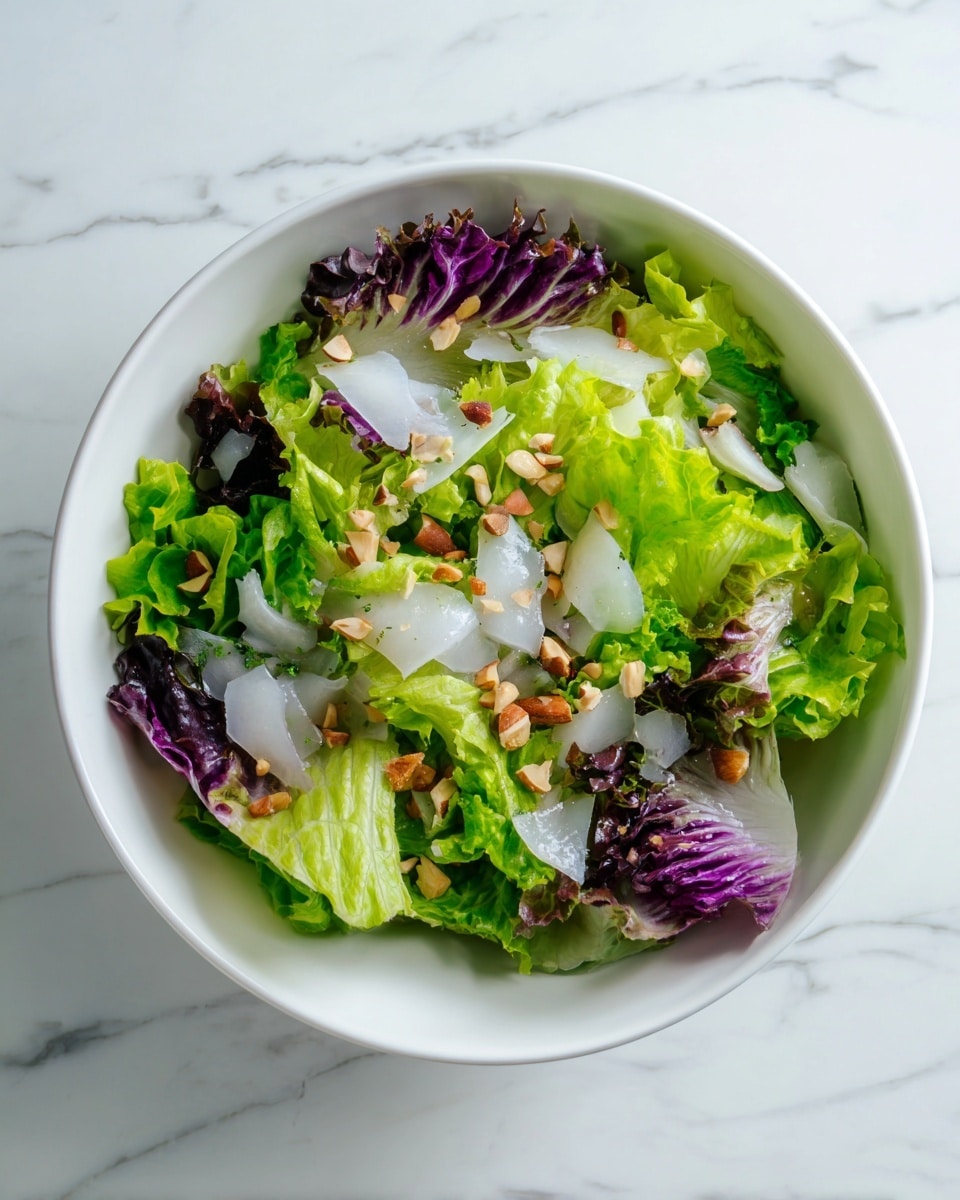 A white bowl filled with a fresh salad made of green and purple lettuce leaves as the base layer. On top, there are thin white strips that look like squid or cuttlefish scattered evenly. Small chopped nuts add texture and are sprinkled across the salad. The bowl is placed on a white marbled surface, showing a clean and bright setting. Photo taken with an iphone --ar 4:5 --v 7