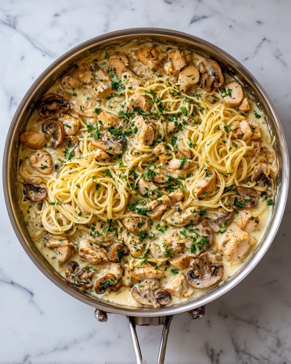 The image shows a stainless steel skillet filled with a creamy pasta dish. The bottom layer has light brown cooked mushrooms with soft textures nestled around the edge of the pan. Above that, there are light yellow cooked spaghetti noodles mixed evenly throughout. There are pieces of pale, cooked chicken scattered on top of and within the noodles. Bright green chopped herbs are sprinkled over the entire dish as a final layer, adding a fresh look. The skillet sits on a white marbled surface. Photo taken with an iphone --ar 4:5 --v 7