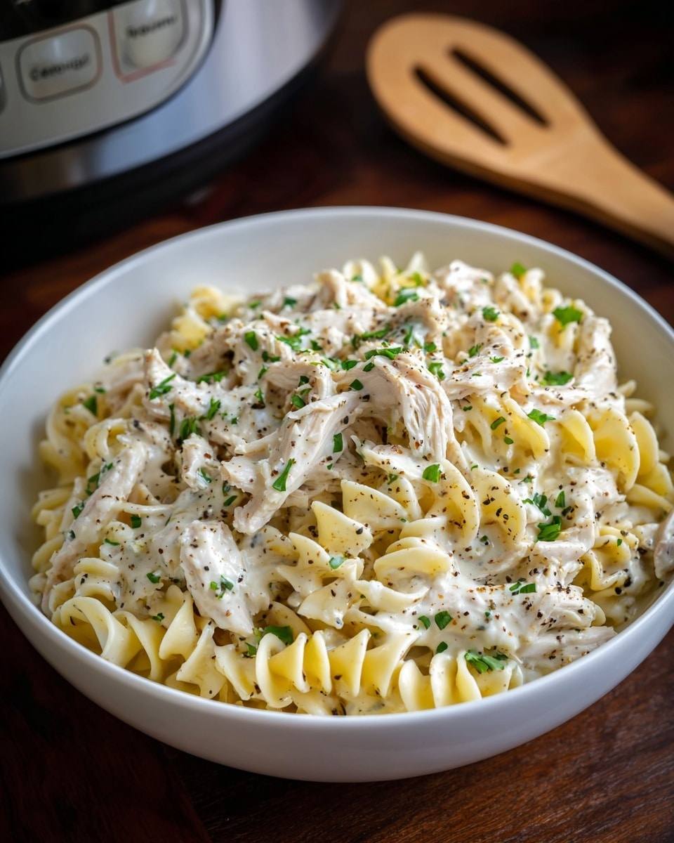 A white round bowl filled with curly egg noodles covered in a creamy white sauce with visible specks of black pepper and chopped green herbs sprinkled on top. There are several pieces of white shredded chicken spread evenly throughout the noodles. The bowl is placed on a dark wooden surface with a wooden slotted spoon near it and a slow cooker in the background. Photo taken with an iphone --ar 4:5 --v 7