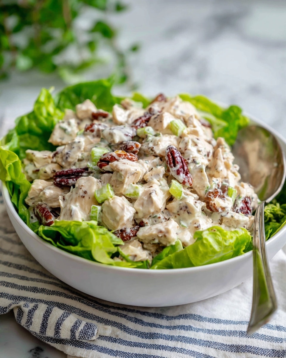 A white bowl filled with a creamy salad that has chunks of light beige chicken mixed with small green celery pieces and dark brown pecans. The salad rests on fresh bright green lettuce leaves lining the bowl. A silver spoon is placed on the right side inside the bowl. The bowl sits on a white and blue striped cloth on a white marbled surface with a blurred green plant in the background. Photo taken with an iphone --ar 4:5 --v 7