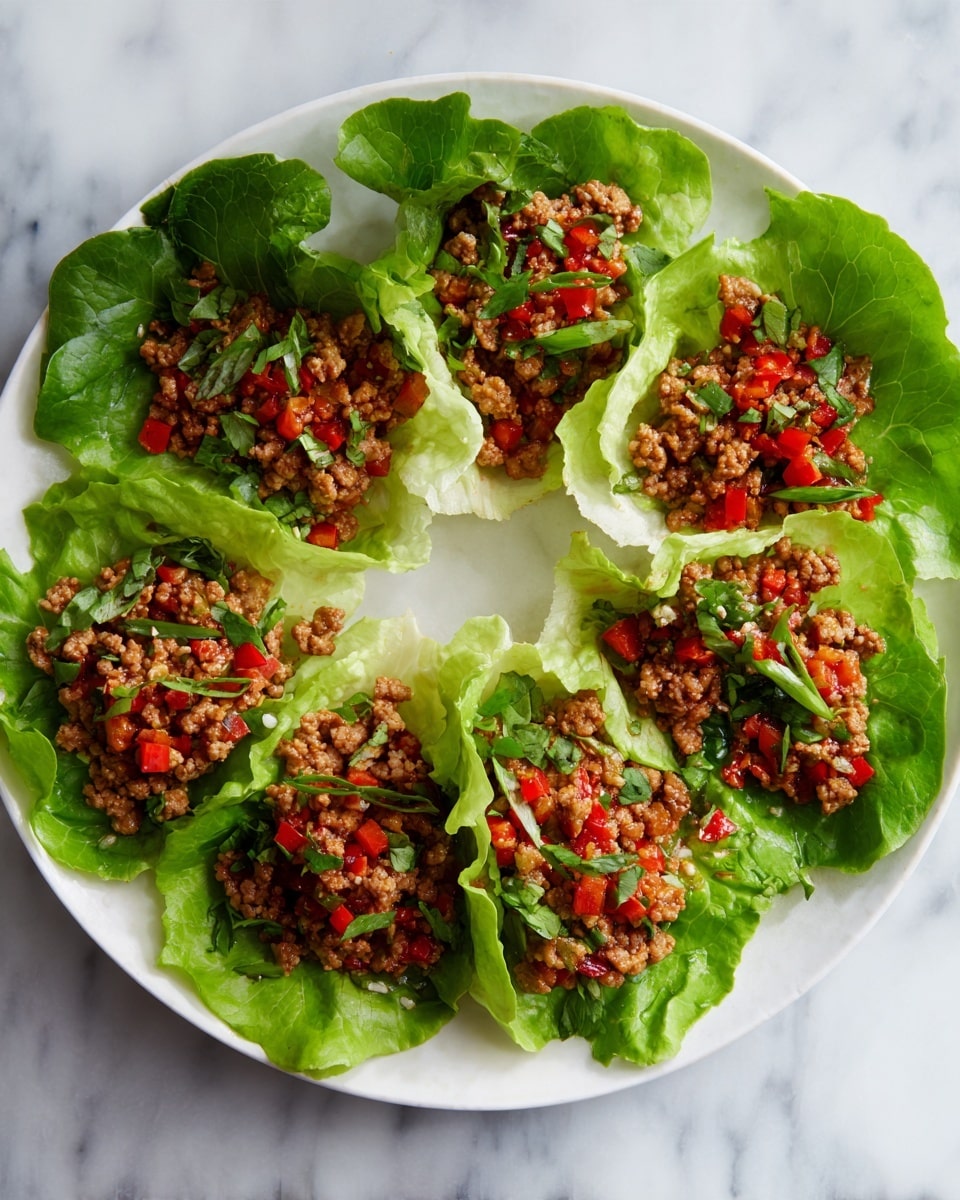 The image shows a white plate filled with six lettuce wraps arranged in a circle. Each wrap has a base layer of light green, crisp lettuce leaves with visible veins and soft texture. On top of the leaves, there is a layer of cooked ground meat mixed with small pieces of bright red diced tomatoes and finely chopped green herbs scattered throughout. The plate sits on a white marbled surface. The overall look is fresh and colorful with a mix of green, red, and brown shades. photo taken with an iphone --ar 4:5 --v 7