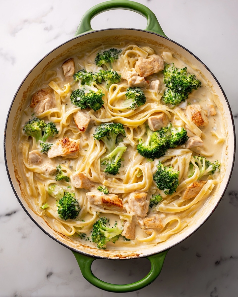 A white cooking pan with a green handle holds a creamy pasta dish with three main layers: the bottom layer is a creamy light beige sauce, the middle layer has long, flat fettuccine noodles in a pale yellow color, and the top layer features bright green broccoli florets and light brown cooked chicken pieces scattered evenly on top. The edges of the pan are lightly stained with the creamy sauce. The whole scene is set on a white marbled surface. photo taken with an iphone --ar 4:5 --v 7