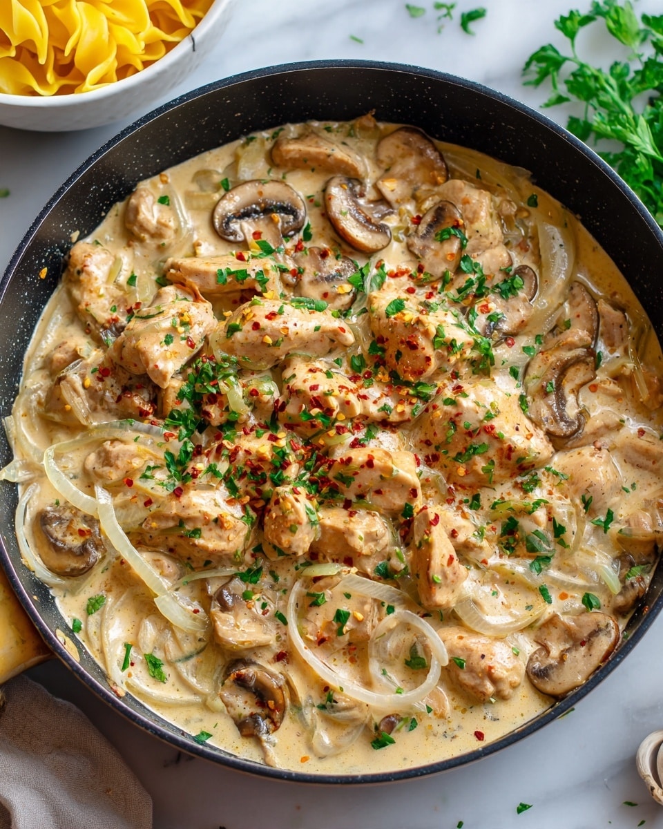 The image shows a black pan filled with creamy sauce and pieces of cooked chicken mixed with sliced mushrooms. The sauce is thick and pale beige, covering thinly sliced onions and garnished with chopped green herbs and a light sprinkle of red spices on top. The pan is placed on a white marbled surface, and part of a white bowl with yellow pasta is visible near the edge. The texture of the sauce looks smooth and rich, with the chicken pieces appearing tender and well-coated in the sauce. Photo taken with an iphone --ar 4:5 --v 7