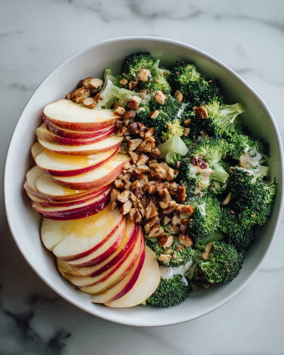 A white bowl holds a fresh salad with several layers. The bottom layer has green broccoli florets with small water droplets showing their fresh and crunchy texture. On top of this, thin slices of red and yellow apple are spread evenly, adding a contrast of smooth and crisp textures with their shiny peel and pale inside. Small pieces of crushed nuts are sprinkled over the apples for a rough, crunchy look. All ingredients sit inside the bowl on a white marbled surface, and the photo is taken with an iphone --ar 4:5 --v 7