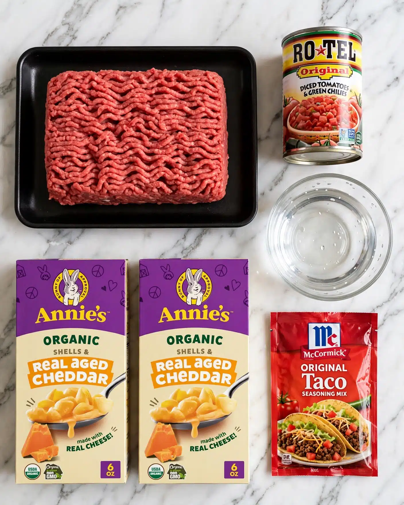 Quick and Easy Taco Mac and Cheese Recipe 4 Square image A black tray holds a block of raw red ground meat with a soft, textured surface full of small ridges, placed on a white marbled wood background. To the right of the meat is a small glass bowl with clear water, and above it is a can of diced tomatoes with green chili, showing bright red tomatoes on a white label with yellow, green, and red stripes. Below the meat tray are two yellow and purple boxes of Annie's Real Aged Cheddar shells and cheese pasta, both showing a spoon with creamy yellow pasta and a block of cheddar cheese on the front. At the bottom right, there is a red packet of McCormick Original Taco seasoning mix, decorated with a picture of a filled taco on the front. photo taken with an iphone --ar 4:5 --v 7