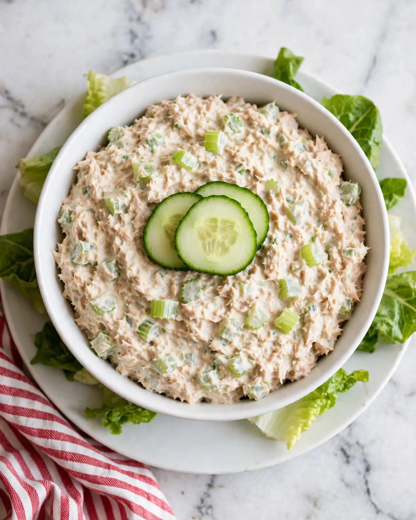 Delicious Cucumber Tuna Salad Recipe 5 Square image The dish shows one bowl filled with a mixture of creamy light beige tuna and light green diced celery evenly spread throughout. Three thin slices of bright green cucumber are placed on top in the center as garnish. The bowl is white and sits on a white plate. Green lettuce leaves peek out from beneath the bowl around the edges. The whole arrangement is placed on a white marbled surface with a red and white striped cloth partly visible. photo taken with an iphone --ar 4:5 --v 7