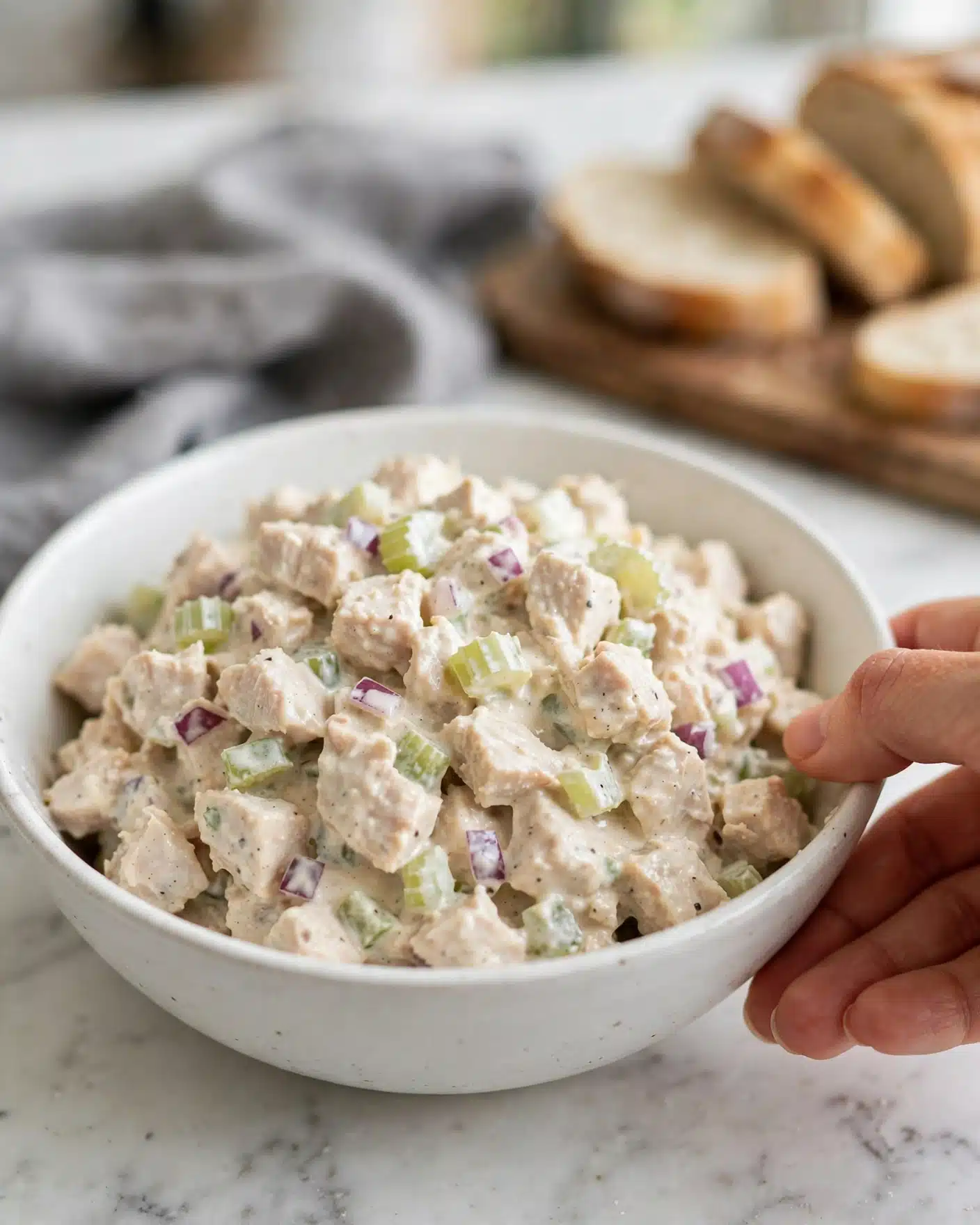Square image A white bowl filled with a creamy salad made of chopped light beige chicken, pale green celery, and small pieces of red onion, all mixed together with a light creamy dressing. The texture looks soft and creamy with bits of crunchy celery and onion visible throughout. The bowl is on a white marbled surface with a gray cloth partially in the background and some sliced bread on a wooden cutting board blurred behind. A woman's hand is just touching the edge of the bowl. Photo taken with an iphone --ar 4:5 --v 7