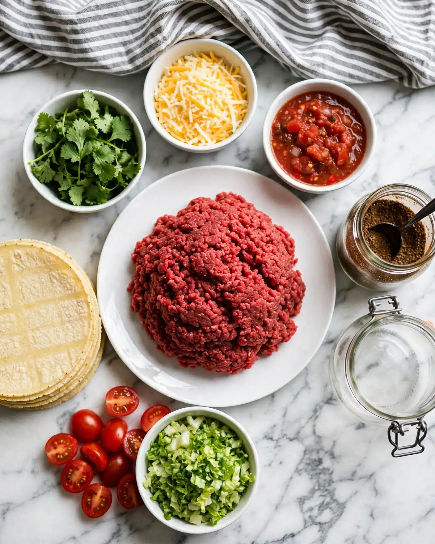 Easy Homemade Ground Meat Tacos Recipe 4 Square image A white plate holds a large mound of raw ground beef, deep red in color, placed in the center on a white marbled surface. To the left, a neat stack of pale yellow corn tortillas lays flat. Surrounding the meat plate in small white bowls are bright green fresh cilantro leaves, shredded yellow and white cheese, and chunky red salsa. Below, halved shiny red cherry tomatoes and a bowl of finely chopped light and dark green lettuce add more color contrast. On the right, a clear glass jar with dark brown spices and a black spoon inside sits next to a jar with a metal clasp. A gray and white striped cloth is casually draped at the top. Photo taken with an iphone --ar 4:5 --v 7