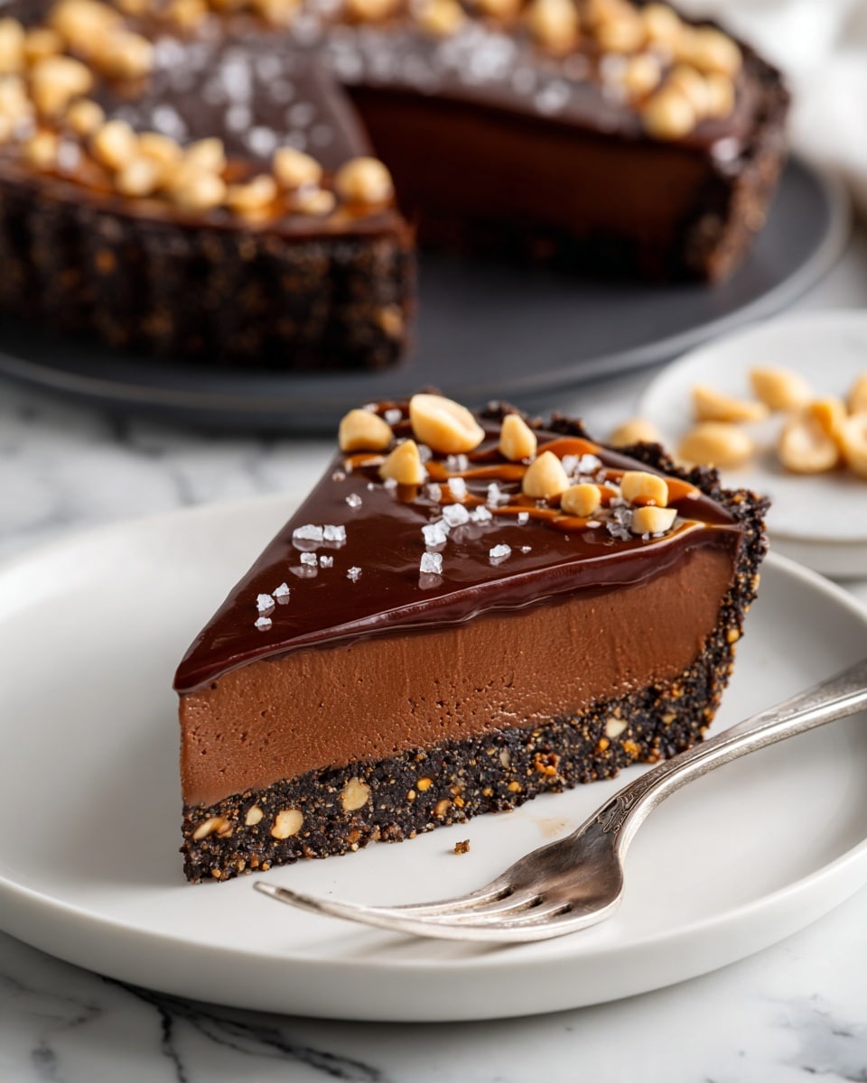 A slice of rich, dark chocolate tart sits on a white plate, showing three layers: a crunchy dark brown crust with visible bits of nuts, a thick, smooth, and creamy chocolate filling in the middle, and a thin glossy chocolate glaze on top. The top layer is decorated with a lighter brown caramel drizzle and scattered whole peanuts, along with small flakes of sea salt. Behind the slice, the whole tart with the same textures and decorations is on a dark plate. The setting is on a white marbled surface. A fork rests next to the slice on the plate photo taken with an iphone --ar 4:5 --v 7