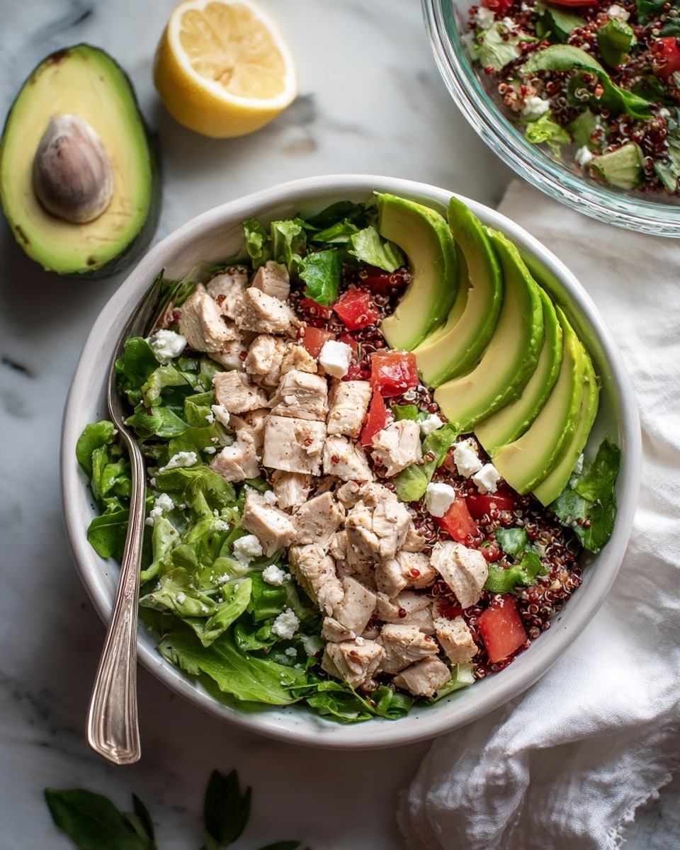 A white bowl filled with a colorful salad layers diced cooked chicken, red quinoa, chopped green lettuce, small pieces of red tomato, and white cheese crumbles, all mixed together. On the right side of the bowl, there are several thick slices of pale green avocado leaning against the salad. A silver fork is placed inside the bowl on the left side. The bowl sits on a white marbled surface along with a half-cut avocado showing its seed on the left and a half lemon with a bright yellow inside near the top. The corner of a glass bowl with more salad is visible at the upper right, resting on a white cloth. Small green leaves are scattered near the bottom, and the lighting is soft and natural. Photo taken with an iphone --ar 4:5 --v 7