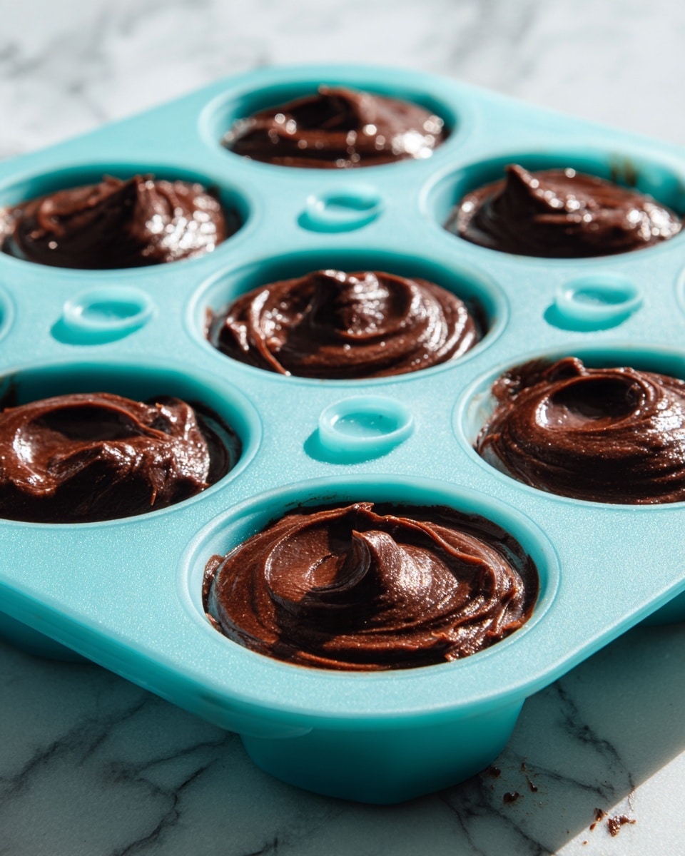 The image shows a turquoise silicone mold with six round cavities, each filled with thick, dark chocolate batter. The batter has a smooth and shiny texture, slightly swirled on the surface. Each cavity has a small turquoise circle in the center that appears to be part of the mold design. The mold is placed on a white marbled surface with some batter spilled around it. The lighting is soft and natural, highlighting the glossy batter and the vibrant color of the mold. Photo taken with an iphone --ar 4:5 --v 7