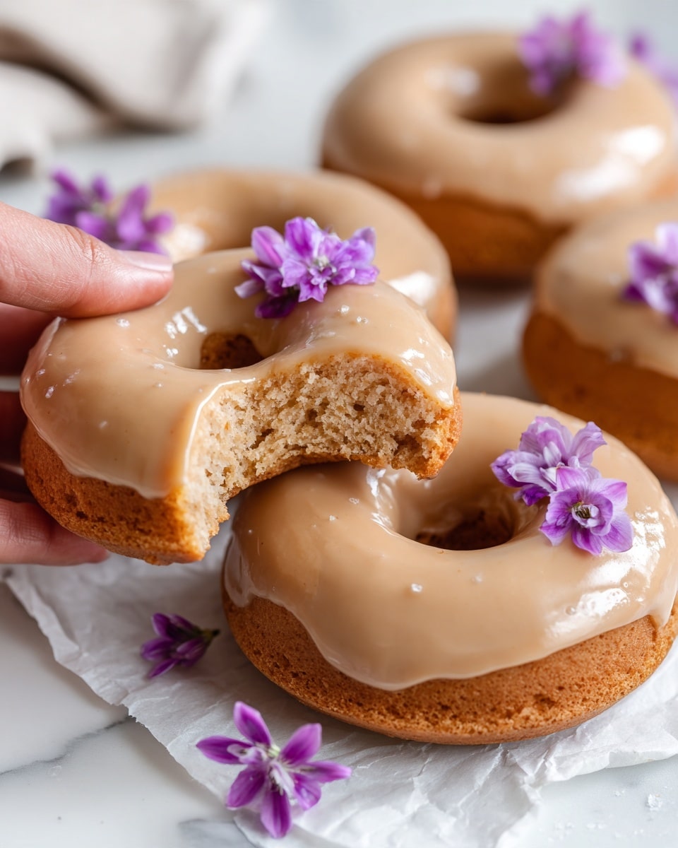 The image shows a close-up of several glazed donuts with a smooth, light brown icing covering the entire top and sides. One donut is missing a bite, revealing a soft, dense inside with a light brown color. The donuts are resting on white parchment paper placed on a white marbled surface. A few small purple edible flowers are delicately placed on some donuts, adding a bright color contrast. A woman's hand is gently holding one donut, showing the texture of the glaze and the soft inside clearly. photo taken with an iphone --ar 4:5 --v 7