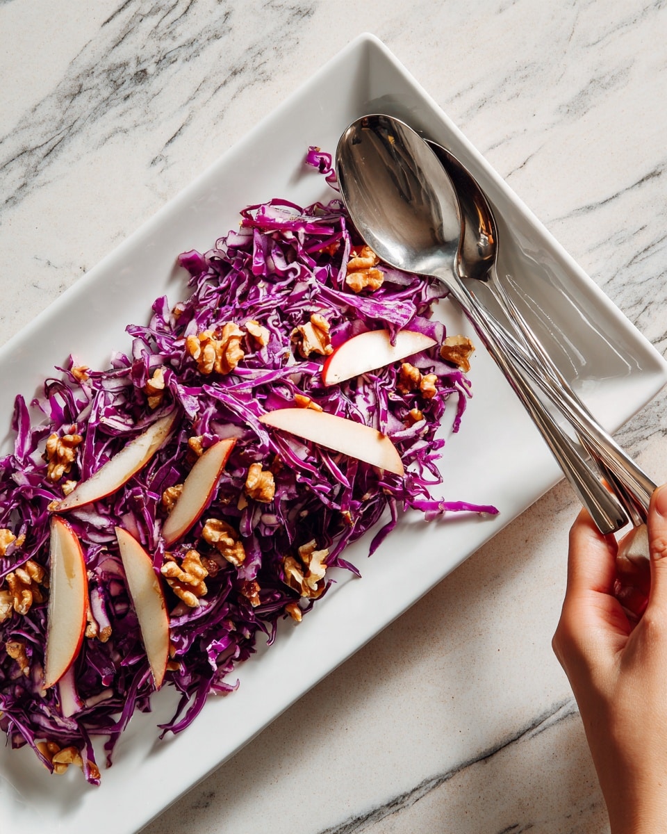 A white rectangular plate holds a colorful salad layered with thinly sliced red cabbage as the base, showing its vibrant purple color and crisp texture. On top, there are evenly spread, thin slices of red-skinned apples with pale cream flesh. Scattered throughout are small pieces of light brown walnuts adding a crunchy texture. The salad looks fresh and slightly glossy, likely from a light dressing. At the edge of the plate, a silver fork and spoon rest on a white marbled surface. A woman's hand is holding the fork near the salad’s top right corner. Photo taken with an iphone --ar 4:5 --v 7