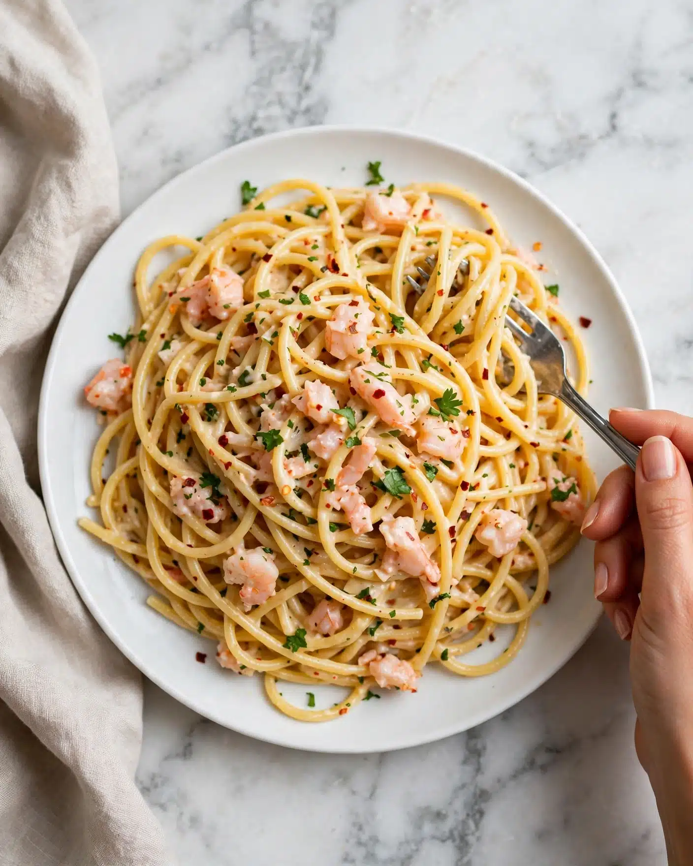 square image A white plate filled with a generous serving of creamy pasta. The pasta strands are long and yellow, covered in a light sauce with small pieces of light pink seafood and bits of green herbs scattered on top. Some red chili flakes add tiny specks of red throughout. A silver fork is tucked in on the right side, with a woman's hand gripping the fork. The plate sits on a white marbled surface with a light beige cloth partly visible on the left side. photo taken with an iphone --ar 4:5 --v 7